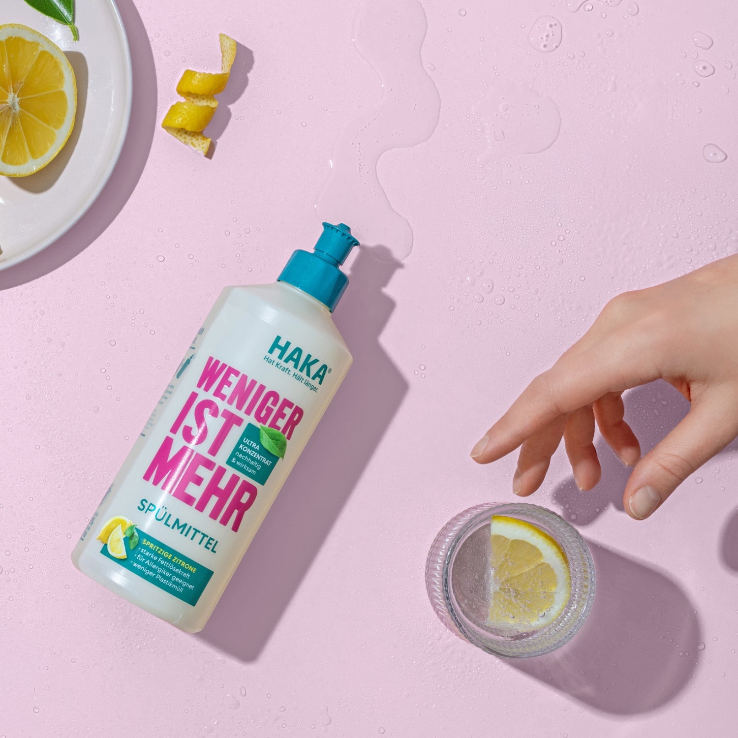 HAKA "Weniger ist Mehr" Spülmittel liegt auf rosa Untergrund neben einer Hand, Zitronenscheiben und einem Glas mit Wasser und Zitrone.